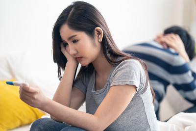 失望的亚洲女人坐在床上时看到了验孕结果。女孩很担心，还没做好当妈妈的准备。男人对抱着未来的孩子感到压