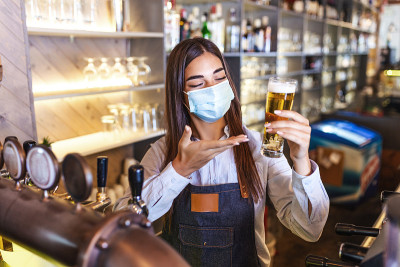 美丽的女调酒师戴着防护面罩，在冠状病毒大流行期间供应生啤酒，背景是摆满酒瓶的架子