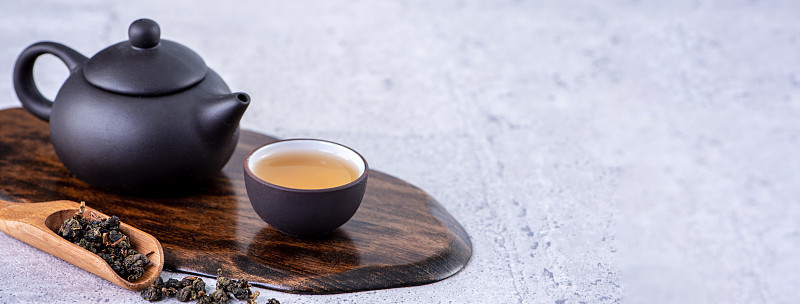 黑色茶壶和茶杯中的热茶，亮灰色水泥背景上的干茶叶，近距离复制空间的设计理念。