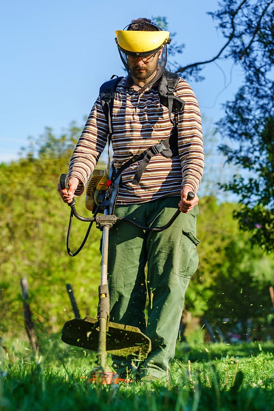 年轻的白人男子，农民，园丁站在地里用线绳修剪，汽油，剪草机割杂草，在地里干活，在阳光明媚的日子里，正