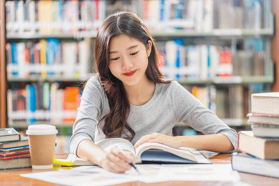 亚洲青年学生穿着休闲服在大学或学院的图书馆里阅读和做作业，书架背景上方的木桌上放着各种书籍和文具，回