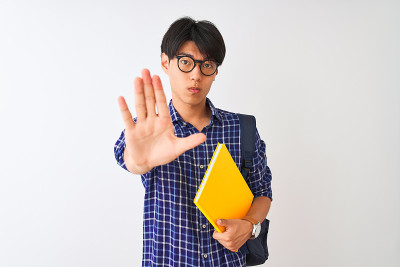 中国学生男子背着背包，拿着笔记本，在孤立的白色背景下，张开手做着停止标志，表情严肃自信，做着防卫姿势