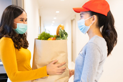 亚洲送人戴着口罩在红色制服处理袋食品，水果，蔬菜给公寓女顾客。新冠肺炎期间的邮差杂货送货服务。预防病