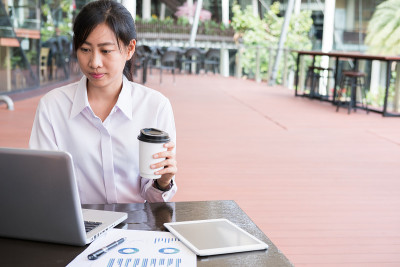 女商人拿着笔记本电脑和财务图表坐在办公大楼外。年轻的亚洲女性在户外分析投资图表。用一次性纸杯装咖啡的