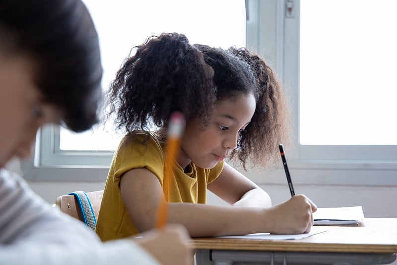 一位非裔美国女学生记笔记在教室里老师讲课。多元化的小学生或孩子专注于学习。回归学校理念