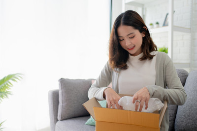 亚洲女人喜欢坐在家里的沙发上。她正在打开一个从网上订购的纸盒。便捷服务的商务交付理念