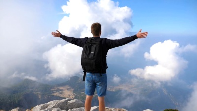 一个面目全非的男孩游客站在山顶上胜利地伸出双臂。在夏季旅行中享受自由的家伙。年轻的男性徒步旅行者，背着背包登上山顶，举起手来。后视图慢动作