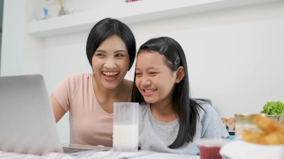 慢动作亚洲母女一起在厨房里用笔记本电脑看视频，面带微笑，喝着牛奶。在家里学习。