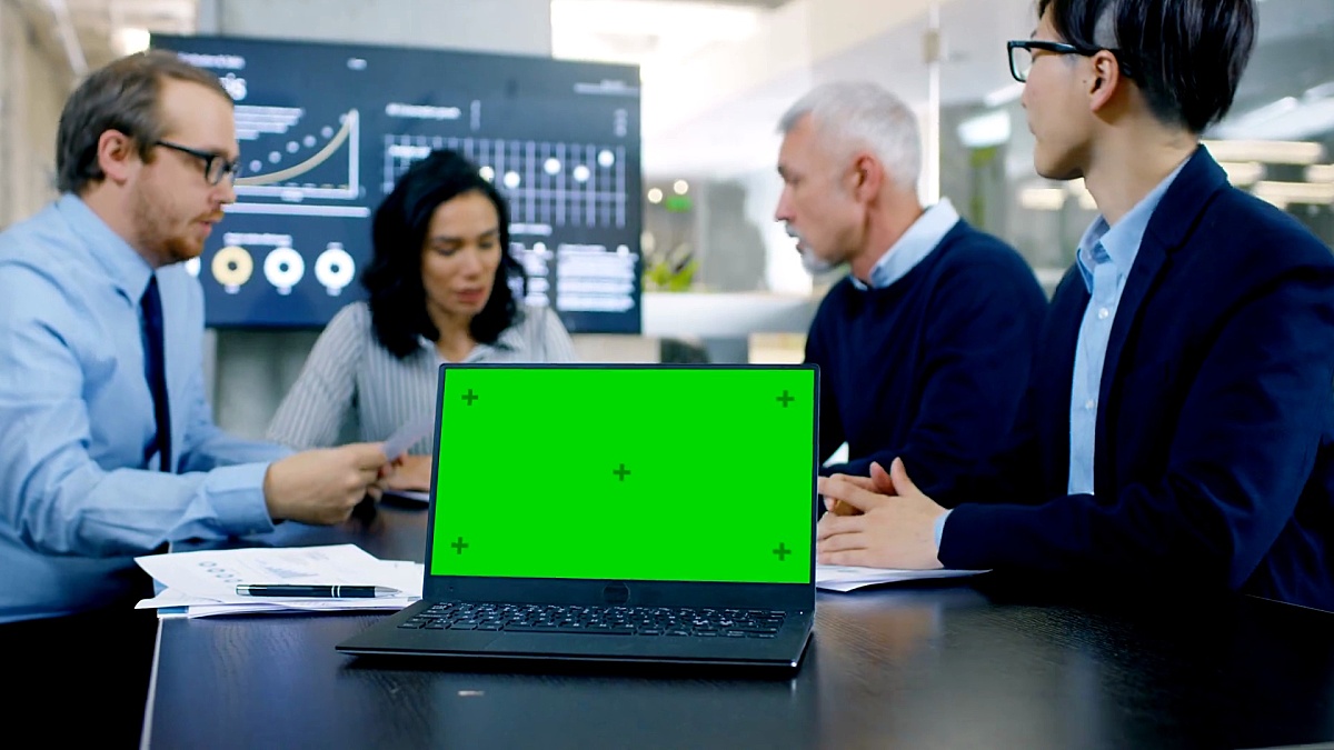 在会议室的笔记本电脑上，会议桌上有绿色色度键屏幕。在背景中，商务人士有重要的讨论。