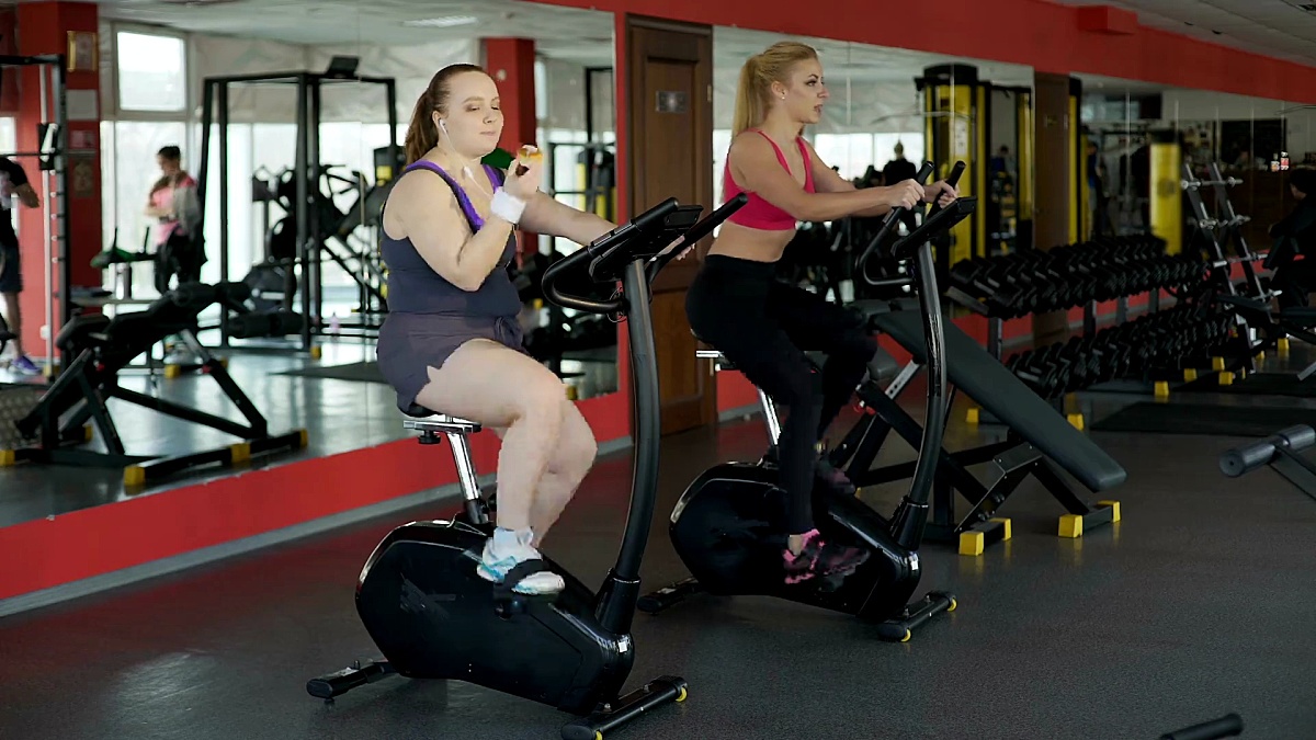 懒惰肥胖的女人在健身房里吃着包子骑着健身自行车