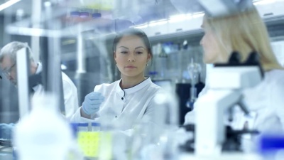 女性研究科学家将通行证滑给她的同事，她的同事在显微镜下观察样品。他们在一个大型研究中心/实验室工作。