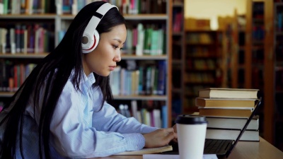 美丽的亚洲女学生戴着耳机坐在图书馆的办公桌前，集中精力在学校项目上工作。年轻女孩在笔记本电脑上打字，喝咖啡，在抄写本上写字