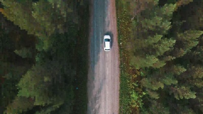 在松树森林的道路上行驶的汽车的空中拍摄。无人驾驶飞机拍摄砾石路和树木，跟随白色汽车
