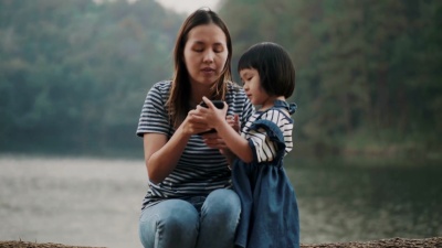 泰国单身母亲带着女儿在绿色自然环境的池塘里用智能手机与父亲正面交谈