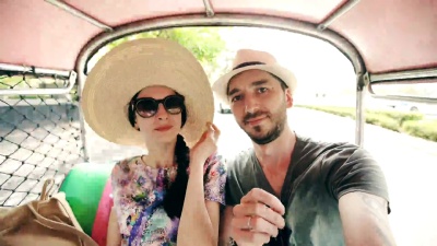 戴着帽子的年轻帅哥和他的女朋友坐在传统的泰国公交车上，在智能手机上自拍。夫妇在蜜月期间去泰国旅行