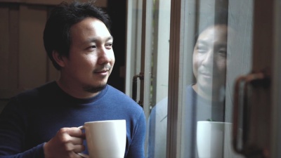  慢动作镜头，亚洲帅哥站在窗玻璃旁，微笑着喝咖啡，从咖啡馆向外望去，生活方式和休闲理念