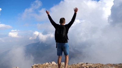 年轻的男性徒步旅行者，背着背包在山边跳跃，胜利地举起双手。男人在夏季旅行中享受自由。背景是多云的天空。后视图慢动作