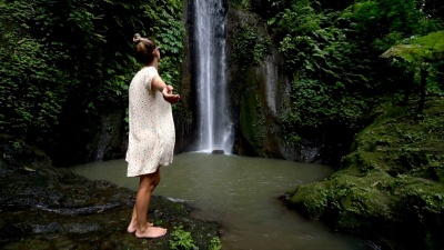 慢动作年轻女子享受在巴厘岛，印度尼西亚，亚洲瀑布下的清新。人们旅行有趣的自然概念。女孩张开双臂