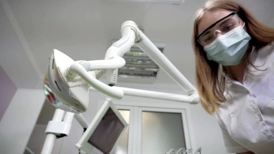 年轻的女牙医戴着面具用工具检查病人，站在病人身上，看着相机，牙医的脸