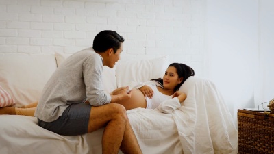 美丽的孕妇和她的丈夫在家里的沙发上在肚子上涂护肤霜，怀孕和家庭观念