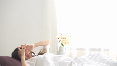 快乐美丽的亚洲年轻女子早上醒来，坐在床上，舒舒服服地躺在舒适的卧室里，透过窗户看。搞笑的亚洲女人醒来后。亚洲女人在伸展和微笑。