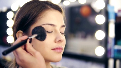 Closeup view of female hands applying facial powder on young woman's skin using special brush. Slowmotion shot