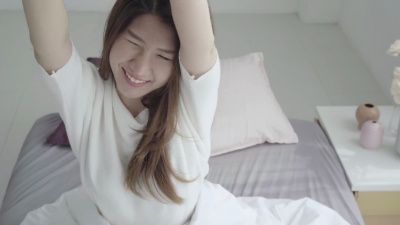 快乐美丽的亚洲年轻女子早上醒来，坐在床上，舒舒服服地躺在舒适的卧室里，透过窗户看。搞笑的亚洲女人醒来后。亚洲女人在伸展和微笑。