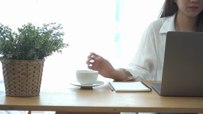 坐在家里的起居室里，坐在笔记本电脑上工作的年轻美丽的微笑女子一边享受着喝热咖啡的乐趣。享受在家的时光。在家庭办公室工作的亚洲女商人。