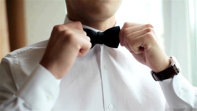 穿衣服的男人近距离拉直领结。衣着考究的年轻人在白色衬衫上调整经典的黑色领结，没有脸，只有微笑。男士之手准备婚礼校正领带和形象