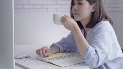 坐在家里的起居室里，坐在笔记本电脑上工作的年轻美丽的微笑女子一边享受着喝热咖啡的乐趣。享受在家的时光。在家庭办公室工作的亚洲女商人。