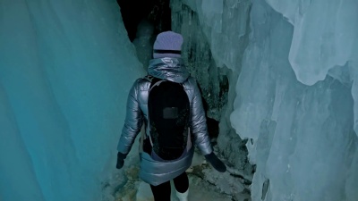 贝加尔湖冰洞上的旅行者。冬岛之旅。女孩背包客走在冰窟里。旅行者看着美丽的冰窟。徒步旅行者穿着银色夹克，背包，LED灯。
