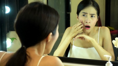 年轻的亚洲女孩有痤疮。少女女性的肖像看着镜子，在浴室里挤粉刺以求美容，皮肤护理生活方式的概念
