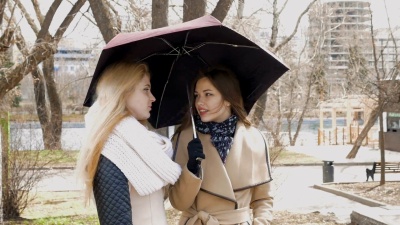 在公园见朋友。下雨时，两个金发女孩和一个深色头发的女孩站在雨伞下。一个年轻人走近他们。他们兴高采烈地打招呼，聊天。