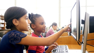 在STEM学校，一名西班牙裔女中学生在电脑上辅导一名非裔美国女小学生