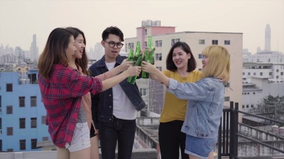 一群年轻的亚洲男女在屋顶的落日城市派对上随着DJ演奏的音乐在空中跳舞和举起手臂。年轻的亚洲女孩和男孩朋友一起喝酒