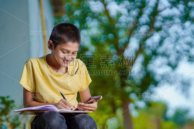 印度一名男孩手持手机，使用手机应用程序进行远程学习，观看在线课程，视频呼叫在应用程序中做笔记，在家学