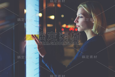 在巴塞罗那，一名年轻女子在用智能公交站选择城市交通时刻表时，触摸着敏感的屏幕，她站在大屏幕前，屏幕上