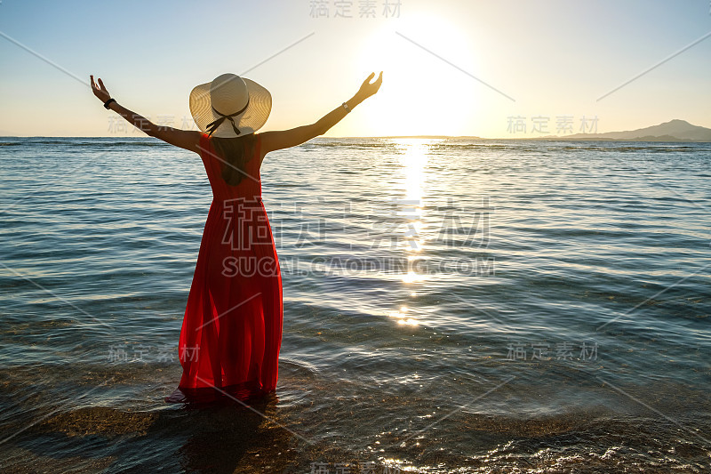 初夏清晨，一名身穿红色长裙、头戴草帽的年轻女子站在海边的海水中，举手欣赏初升的太阳。