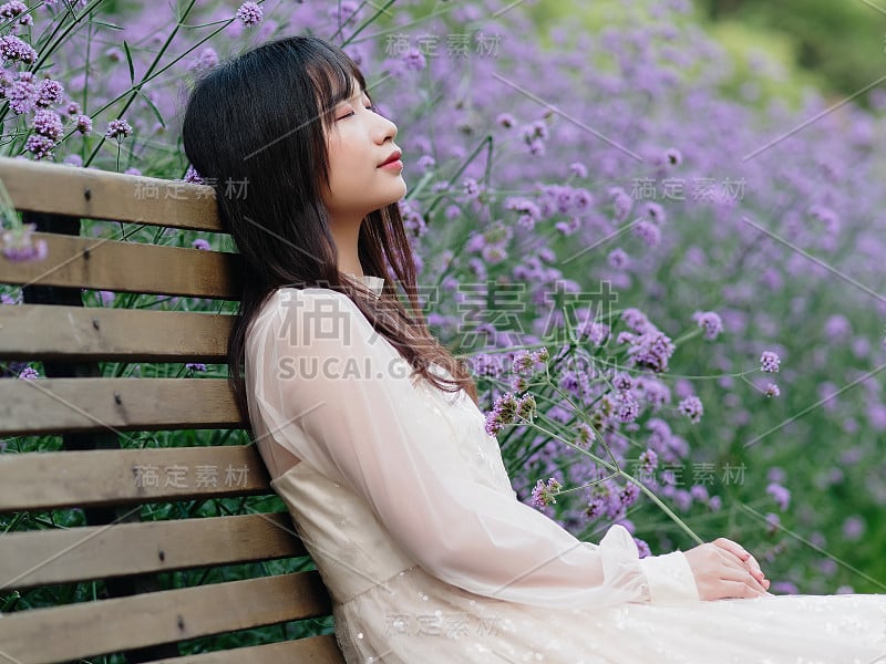 美丽的女人在白色的裙子摆姿势在紫色的马鞭草花田，迷人的中国女孩与黑色的长发享受她的休闲时间户外。