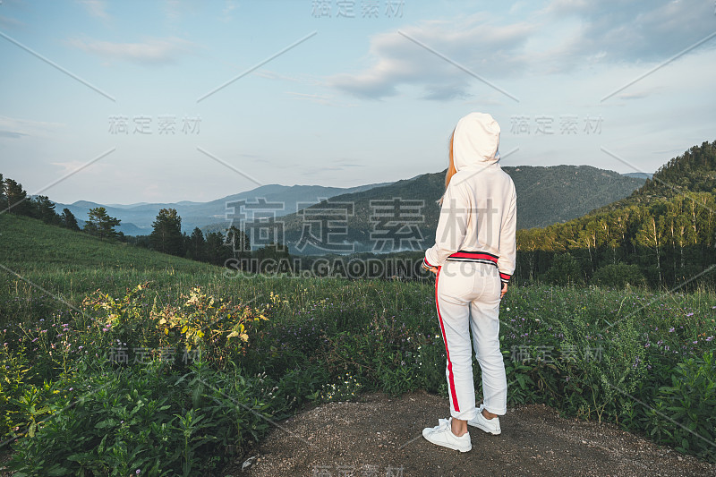 傍晚时分，一个穿着白色运动服的年轻女孩凝视着远处美丽的群山。爬山。去山上的旅程。户外活动。