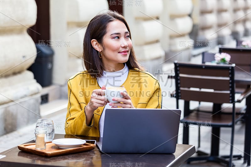 年轻的亚洲商务女性坐在桌子上，在笔记本上记笔记。桌上放着笔记本电脑、智能手机和一杯咖啡。电脑屏幕上的