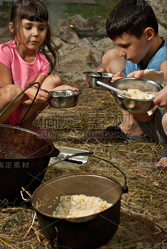 孩子们在露营时吃东西。锅里的食物是用火煮的。女孩和男孩拿着面具。孩子们把食物扔在盘子里。