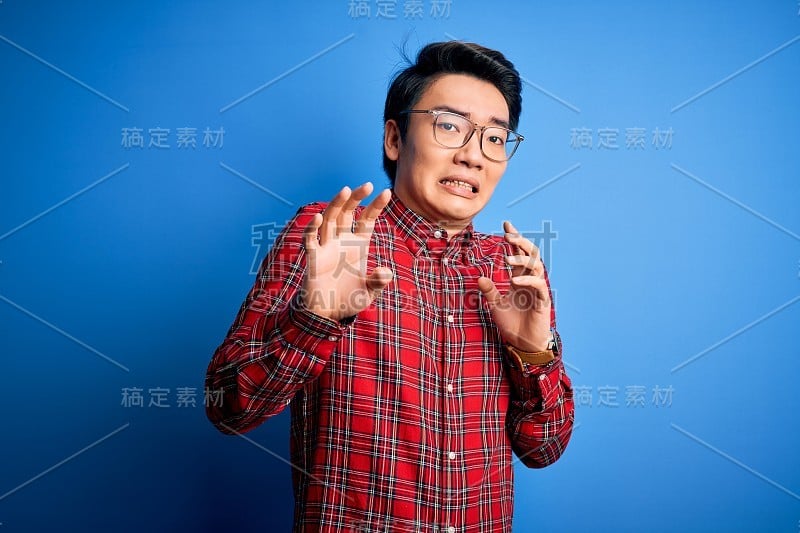 年轻英俊的中国男子穿着休闲衬衫和眼镜在蓝色的背景下表现出厌恶的表情，不高兴和恐惧做厌恶的脸因为厌恶的