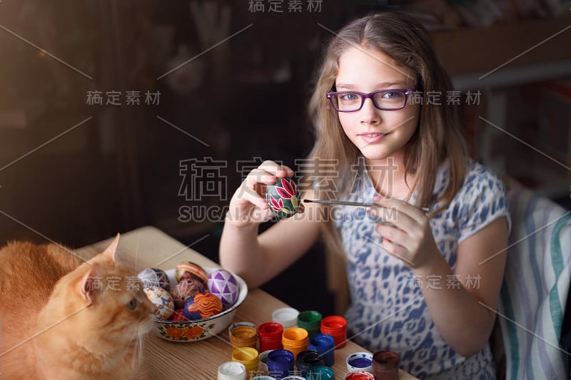 十几岁的女孩画复活节彩蛋，她的姜黄色的猫躺在桌子上。