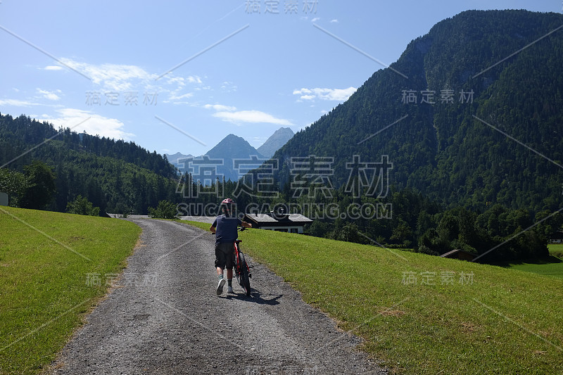 小男孩穿着短裤和自行车头盔推着他的自行车上山在一条土路在他活跃的暑假在奥地利山区。