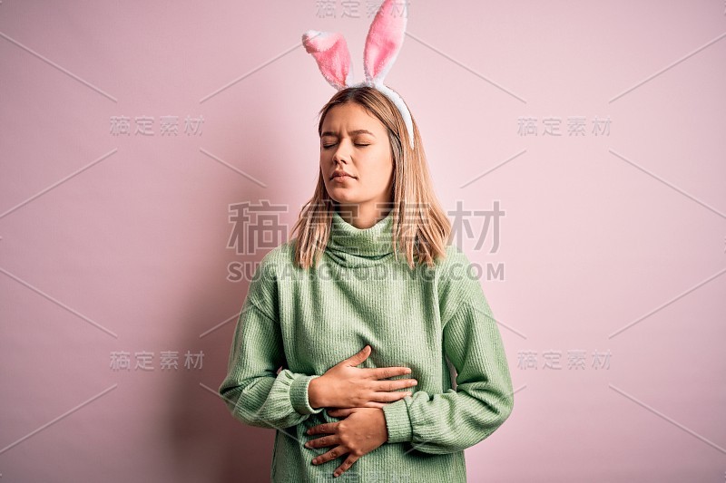 年轻美丽的女子戴着复活节兔子耳朵站在孤立的粉红色背景与手胃，因为消化不良，痛苦的疾病感觉不舒服。疼痛