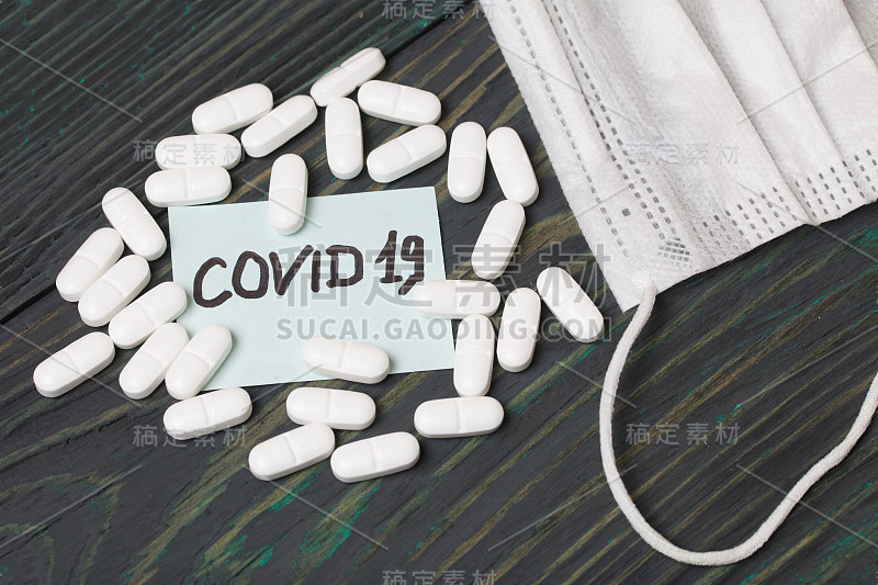 贴有COVID-19字样的贴纸。附近有几粒白色药丸和一个防护面具。躺在刷过的松木板上。