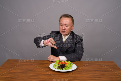 工作室拍摄的成熟的亚洲商人坐在木桌子上，以灰色的背景