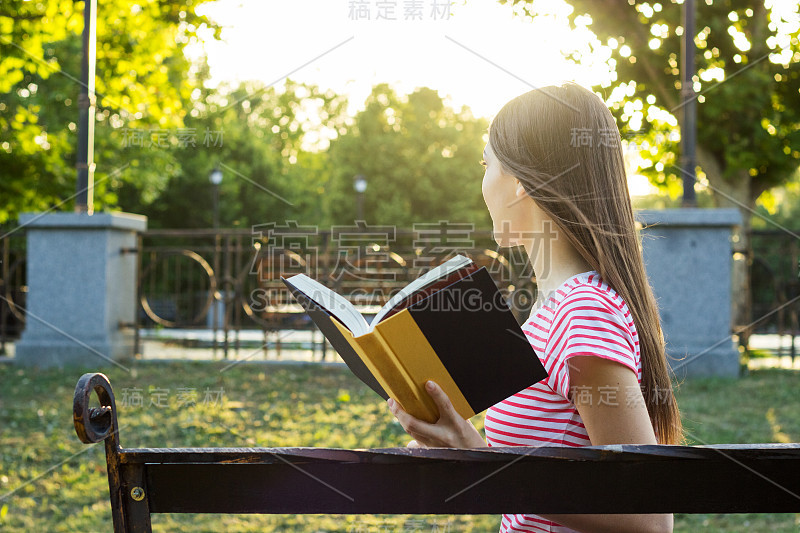 一个迷人的年轻女孩在夏天阳光明媚的一天在公园里看书的侧视图。