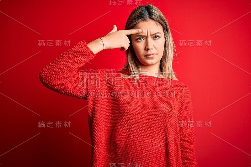 年轻美丽的金发女人穿着休闲毛衣在红色孤立的背景指向不愉快的丘疹在前额，丑陋的感染黑头。痤疮和皮肤问题